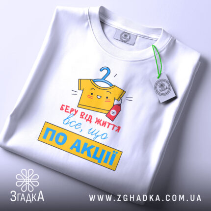Футболка все, що по акції. Біла футболка, складена на столі, з видимим принтом. Фон світло-сірий, принт кольоровий, з жовтою футболкою на вішалці та червоною пляшечкою, текст червоний і жовтий, з зеленим ярликом.