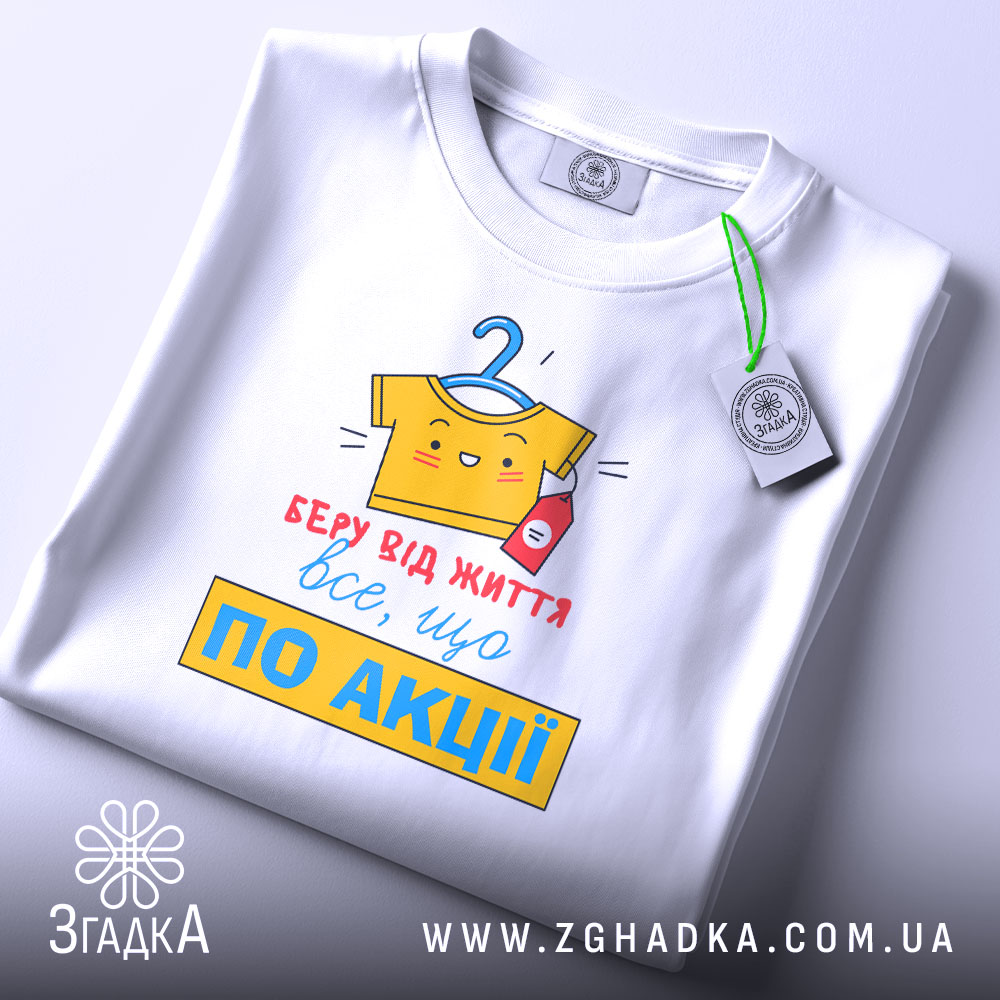 Футболка все, що по акції. Біла футболка, складена на столі, з видимим принтом. Фон світло-сірий, принт кольоровий, з жовтою футболкою на вішалці та червоною пляшечкою, текст червоний і жовтий, з зеленим ярликом.