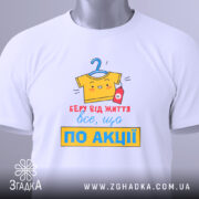 Футболка все, що по акції. Біла футболка, складена на столі, з видимим принтом. Фон світло-сірий, принт кольоровий, з жовтою футболкою на вішалці та червоною пляшечкою, текст червоний і жовтий, з зеленим ярликом.