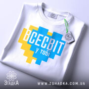 Футболка всесвіт у тобі, біла, складена на столі, фон світло-сірий, принт у формі серця з жовтими та блакитними квадратами, текст "ВСЕСВІТ у тобі" великими буквами, на етикетці зелена стрічка.