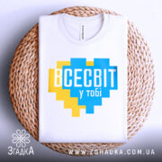 Футболка всесвіт у тобі, біла, складена на плетеній тарілці, фон світло-сірий, принт у формі серця з жовтими та блакитними квадратами, текст "ВСЕСВІТ у тобі" великими буквами.