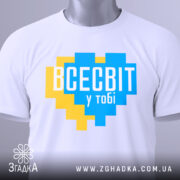 Футболка всесвіт у тобі, біла, складена на столі, фон світло-сірий, принт у формі серця з жовтими та блакитними квадратами, текст "ВСЕСВІТ у тобі" великими буквами, на етикетці зелена стрічка.