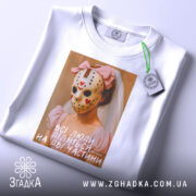 Футболка всі люди діляться, біла, зображена з верхнього ракурсу, на фоні світло-сірого градієнта, принт з дівчиною в рожевій сукні та масці, з червоними сердечками, підпис на принті: 'всі люди діляться на дві частини', з етикеткою на рукаві.