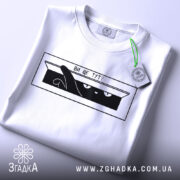 Футболка ви ще тут, білого кольору, складена на поверхні, фон світло-сірий, принт чорного кольору з зображенням кота, який виглядає з коробки, текст "ВИ ЩЕ ТУТ?", на футболці є етикетка з зеленим шнурком.