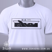 Футболка ви ще тут, білого кольору, складена на поверхні, фон світло-сірий, принт чорного кольору з зображенням кота, який виглядає з коробки, текст "ВИ ЩЕ ТУТ?", на футболці є етикетка з зеленим шнурком.