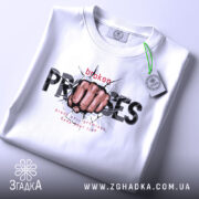 Футболка вибух емоцій. Біла футболка, складена на столі, з принтом кулака, що пробиває стіну, текстом 'broken PROMISES'. На футболці є етикетка з зеленим шнурком. Фон світлий, акцент на деталі принта.