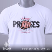 Футболка вибух емоцій. Біла футболка, складена на столі, з принтом кулака, що пробиває стіну, текстом 'broken PROMISES'. На футболці є етикетка з зеленим шнурком. Фон світлий, акцент на деталі принта.