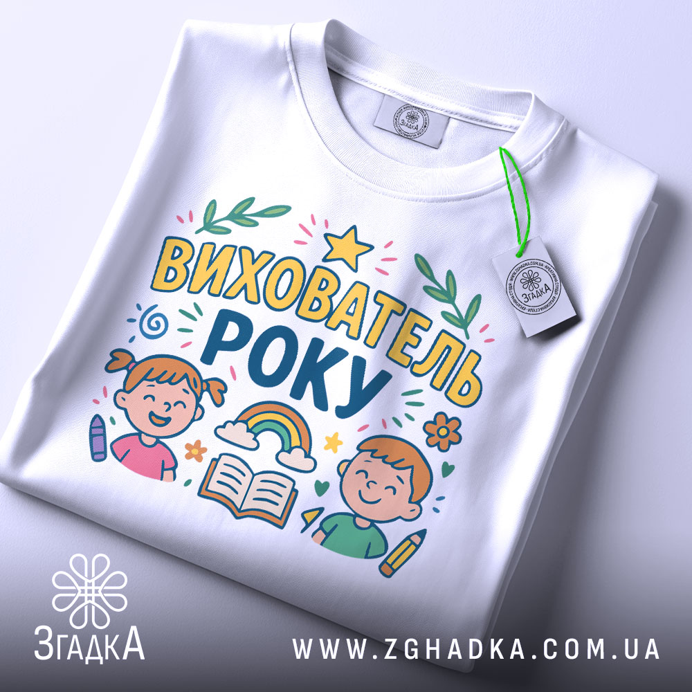 Футболка вихователь року, біла, зображена зверху, на ній видно принт з малюнками дітей, книжки та веселки, етикетка з зеленим шнурком прикріплена до коміра.