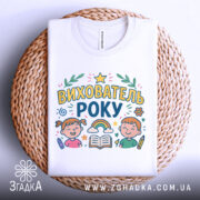 Футболка вихователь року, біла, складена на плетеній основі, принт з малюнками дітей, книжки та веселки, фон світлий.