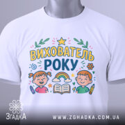 Футболка вихователь року, біла, зображена зверху, на ній видно принт з малюнками дітей, книжки та веселки, етикетка з зеленим шнурком прикріплена до коміра.