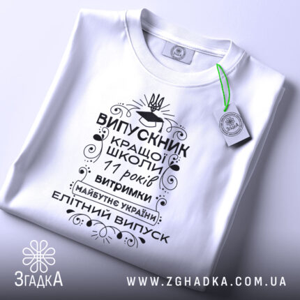 Футболка випускник кращої школи, білого кольору, зображена зверху, на плоскій поверхні, фон світло-сірий, принт чорного кольору з написом та елементами декору, на комірці видна етикетка.