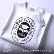 Футболка випускник витримка, біла, складена на поверхні, фон світло-сірий, принт чорного кольору з написом 'ВИПУСКНИК' та зображенням диплома, на футболці є етикетка з зеленим шнурком.