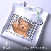 Футболка вирвався з системи, біла, складена на столі, фон світло-сірий, принт з ведмедиком, що виглядає з-під смужок, текст під принтом українською мовою, з етикеткою на шнурку.