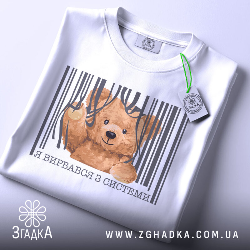 Футболка вирвався з системи, біла, складена на столі, фон світло-сірий, принт з ведмедиком, що виглядає з-під смужок, текст під принтом українською мовою, з етикеткою на шнурку.
