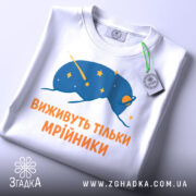 Футболка виживуть тільки мрійники. Біла футболка, зображена зверху, з принтом синього кольору, що зображає космічну істоту з зірками. Текст оранжевого кольору розташований під малюнком. На футболці видно етикетку з зеленим шнурком.