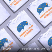 Футболка виживуть тільки мрійники. Біла футболка з принтом, на якому зображено синій малюнок, що нагадує космічну істоту, оточену зірками. Текст на футболці виконаний оранжевим кольором. Фон нейтральний, з легким градієнтом.