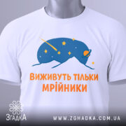 Футболка виживуть тільки мрійники. Біла футболка, зображена зверху, з принтом синього кольору, що зображає космічну істоту з зірками. Текст оранжевого кольору розташований під малюнком. На футболці видно етикетку з зеленим шнурком.