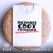 Футболка визнаю свої помилки, білого кольору, складена на плоскій поверхні, фон світлий, принт чорного кольору з червоним акцентом, текст розташований по центру, на комірці видна етикетка.