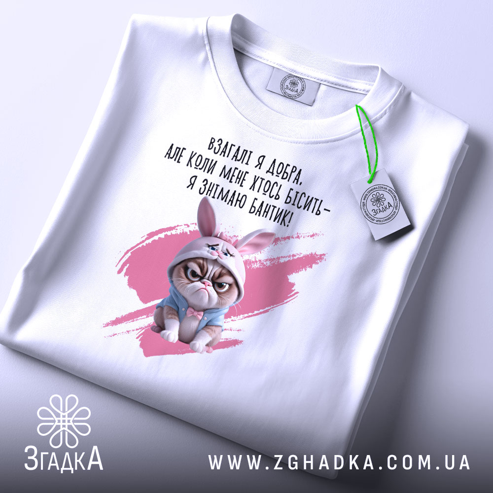 Футболка взагалі я добра. Білий колір, зображена в горизонтальному положенні, фон світло-сірий. Принт з кішкою в зайцевому капюшоні та текстом на українській мові, з зеленим ярликом.