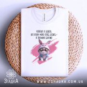 Футболка взагалі я добра. Білий колір, зображена в горизонтальному положенні на плетеній основі, фон світлий. Принт з кішкою в зайцевому капюшоні та текстом на українській мові.