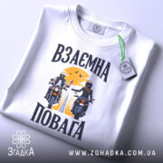 Футболка взаємна повага байкери, біла, складена на столі, фон світло-сірий, принт з двома байкерами, один з них піднімає руку, на фоні жовте сонце і дорога, на комірці є етикетка.