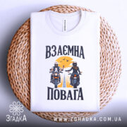 Футболка взаємна повага байкери, біла, складена на плетеній кошику, фон світло-сірий, принт з двома байкерами, один з них піднімає руку, на фоні жовте сонце і дорога.