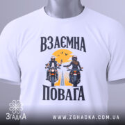 Футболка взаємна повага байкери, біла, складена на столі, фон світло-сірий, принт з двома байкерами, один з них піднімає руку, на фоні жовте сонце і дорога, на комірці є етикетка.