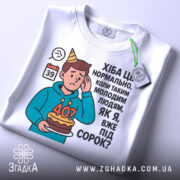 Футболка вже під 40, білого кольору, зображена зверху, на плоскій поверхні, фон світло-сірий, принт з ілюстрацією хлопця з тортом і текстом про вік, на футболці є етикетка.