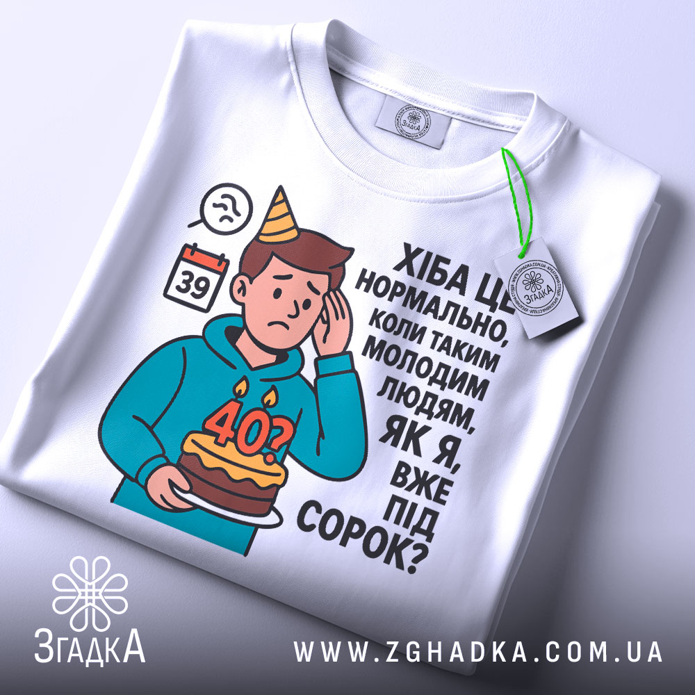 Футболка вже під 40, білого кольору, зображена зверху, на плоскій поверхні, фон світло-сірий, принт з ілюстрацією хлопця з тортом і текстом про вік, на футболці є етикетка.