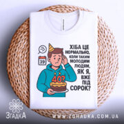 Футболка вже під 40, білого кольору, зображена на плоскій поверхні, фон плетений, принт з ілюстрацією хлопця з тортом і текстом про вік.