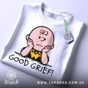 Футболка з Чарлі білого кольору, складена на столі, з принтом Чарлі Брауна, який сидить з опущеними руками, підписом 'GOOD GRIEF!', на етикетці видно логотип бренду, фон світло-сірий.