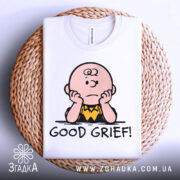 Футболка з Чарлі білого кольору, складена на плетеному кошику, з принтом Чарлі Брауна, який сидить з опущеними руками, підписом 'GOOD GRIEF!', фон світло-сірий.