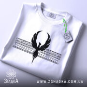 Футболка з феніксом білого кольору, складена на поверхні, фон світло-сірий, принт чорного кольору з феніксом та геометричними елементами, на комірці видна етикетка.