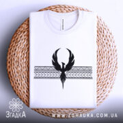 Футболка з феніксом білого кольору, складена на плетеній кошику, фон світло-сірий, принт чорного кольору з феніксом та геометричними елементами, видна етикетка на комірці.