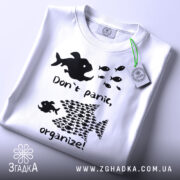 Футболка з філософією: складена біла футболка з чорним принтом, на якому зображені риби та текст "Don't panic, organize!". Фон світлий, на футболці є етикетка з зеленим шнурком.