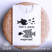 Футболка з філософією: складена біла футболка на плетеній основі, з чорним принтом, що містить риб і текст "Don't panic, organize!". Фон світлий, футболка має круглий виріз.