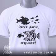 Футболка з філософією: складена біла футболка з чорним принтом, на якому зображені риби та текст "Don't panic, organize!". Фон світлий, на футболці є етикетка з зеленим шнурком.