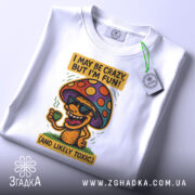 Футболка з грибом, білого кольору, зображена зверху, складена на поверхні, фон світлий, принт з грибом у окулярах, що тримає чашку, з текстом 'I MAY BE CRAZY, BUT I'M FUN!' і 'AND LIKELY TOXIC', з етикеткою на комірці.