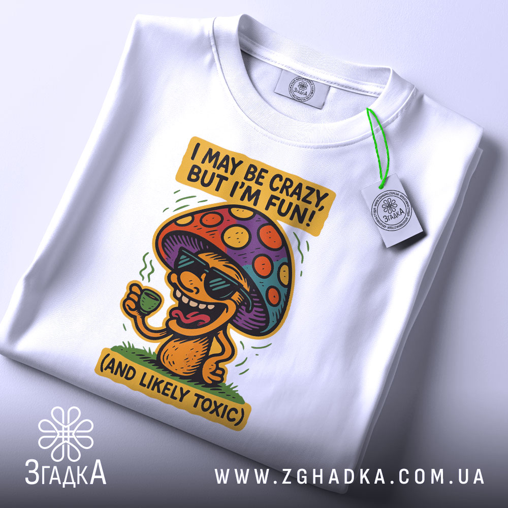 Футболка з грибом, білого кольору, зображена зверху, складена на поверхні, фон світлий, принт з грибом у окулярах, що тримає чашку, з текстом 'I MAY BE CRAZY, BUT I'M FUN!' і 'AND LIKELY TOXIC', з етикеткою на комірці.