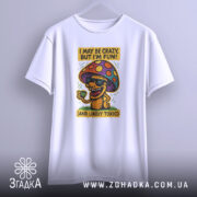 Футболка з грибом, білого кольору, зображена з фронтального ракурсу, підвішена на вішалці, фон нейтральний, принт з веселим грибом у окулярах, що тримає чашку, з текстом 'I MAY BE CRAZY, BUT I'M FUN!' і 'AND LIKELY TOXIC'.