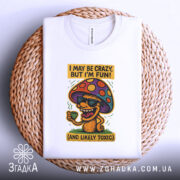 Футболка з грибом, білого кольору, складена на плетеній кошику, фон світлий, принт з грибом у окулярах, що тримає чашку, з текстом 'I MAY BE CRAZY, BUT I'M FUN!' і 'AND LIKELY TOXIC', видно етикетку на комірці.