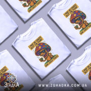 Футболка з грибом, білого кольору, зображена з фронтального ракурсу на манекені, фон нейтральний, принт з веселим грибом у окулярах, що тримає чашку, з текстом 'I MAY BE CRAZY, BUT I'M FUN!' і 'AND LIKELY TOXIC'.