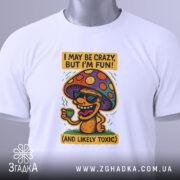 Футболка з грибом, білого кольору, зображена зверху, складена на поверхні, фон світлий, принт з грибом у окулярах, що тримає чашку, з текстом 'I MAY BE CRAZY, BUT I'M FUN!' і 'AND LIKELY TOXIC', з етикеткою на комірці.
