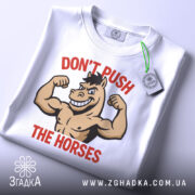 Футболка з конем білого кольору, зображена зверху, на ній видно етикетку, фон світло-сірий, принт з конем, що показує м'язи, з написом 'DON'T PUSH THE HORSES' червоного кольору.