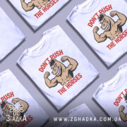 Футболка з конем білого кольору, зображена з фронтального ракурсу на манекені, фон світло-сірий, принт з веселим конем, що демонструє м'язи, з написом 'DON'T PUSH THE HORSES' червоного кольору.