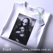 Футболка з Моною Лізою, білого кольору, складена на поверхні, фон світло-сірий, принт зображає Мону Лізу з інопланетянином, на футболці є етикетка з зеленим шнурком.