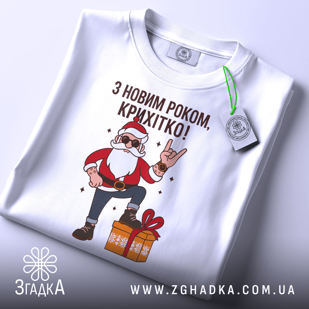 Футболка з Новим Роком, біла, складена на столі, фон світло-сірий, принт з дідом Морозом у червоному костюмі, який піднімає руку в жесті "рок", з зеленим ярликом.