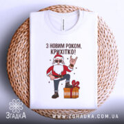 Футболка з Новим Роком, біла, складена на плетеній кошику, фон світло-сірий, принт з дідом Морозом у червоному костюмі, який піднімає руку в жесті "рок", поряд з подарунком.
