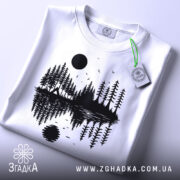 Футболка з природою, біла, складена на поверхні, фон світло-сірий, принт з чорними деревами, озером і сонцем, на футболці є етикетка з зеленим шнурком.