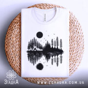 Футболка з природою, біла, складена на плетеній кошику, фон світло-сірий, принт з чорними деревами, озером і сонцем, деталі принта включають відображення в воді.